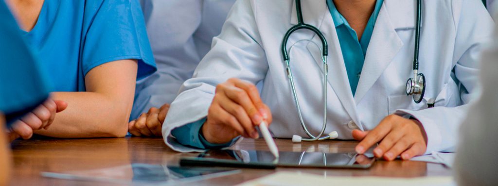 Médica de jaleco usa um tablet, com estetoscópio ao pescoço, em uma reunião hospitalar. O que é Pesquisa Clínica? Entenda a Importância, as Etapas e os Impactos desses Estudos.