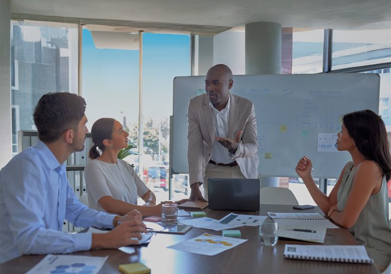 Equipe em reunião sobre tecnologia em gestão de cooperativas, com um homem apresentando em quadro branco e documentos sobre a mesa.