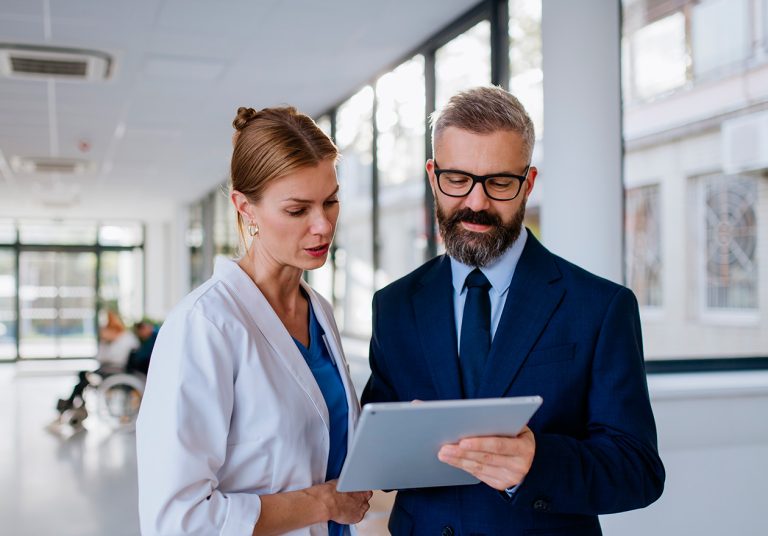 Médica e profissional de terno analisando tablet em corredor de hospital, discutindo dados sobre auditoria em saúde.