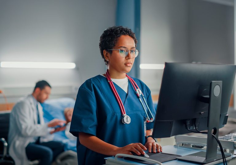 Médica usando computador em hospital analisa informações sobre codificação e análise de informações em saúde na metodologia DRG. Ao fundo, um médico conversa com seu paciente.