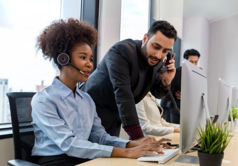 Uma mulher com headset atende no computador e um homem atende o telefone ao lado dela. Curso: A Importância do Atendimento para Otimizar os Resultados.