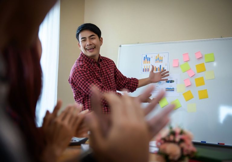 Um homem gesticula em frente a um quadro com post-its coloridos, enquanto pessoas aplaudem. Curso: Aplicando o 5S.