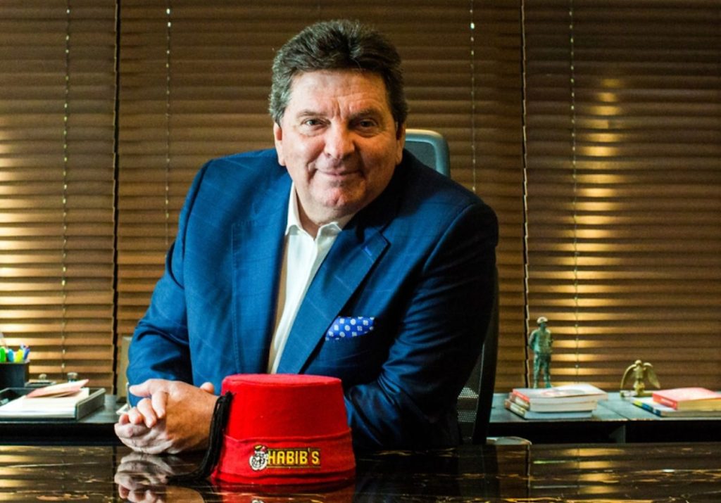 Fundador do Habib's de terno azul sorri em um escritório, com um chapéu vermelho do Habib's na mesa. Curso: Aprenda com o Fundador do Habib's.