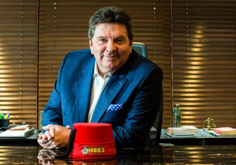 Fundador do Habib's de terno azul sorri em um escritório, com um chapéu vermelho do Habib's na mesa. Curso: Aprenda com o Fundador do Habib's.