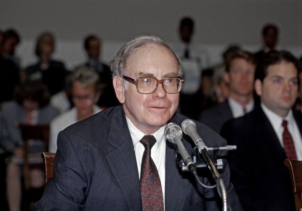 Warren Buffet de terno fala ao microfone em um evento com várias pessoas ao fundo. Curso: Aprenda com Warren Buffett.