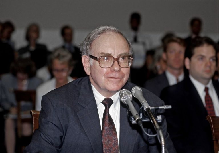 Warren Buffet de terno fala ao microfone em um evento com várias pessoas ao fundo. Curso: Aprenda com Warren Buffett.