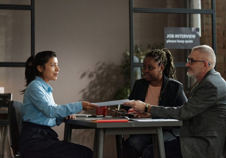 Três pessoas em uma entrevista, com uma mulher entregando um currículo em uma sala de reunião. Curso: Apresentação de Currículo.
