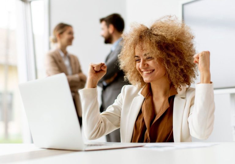 Uma mulher sorridente levanta os braços em frente a um laptop, dois colegas estão ao fundo. Curso: As 10 Competências mais Importantes para a Carreira.