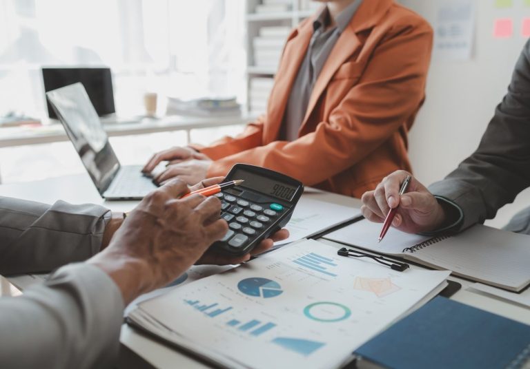 Pessoas em reunião analisam gráficos e usam calculadora sobre uma mesa com laptop. Curso: Aspectos Contábeis e Societários de uma Cooperativa.