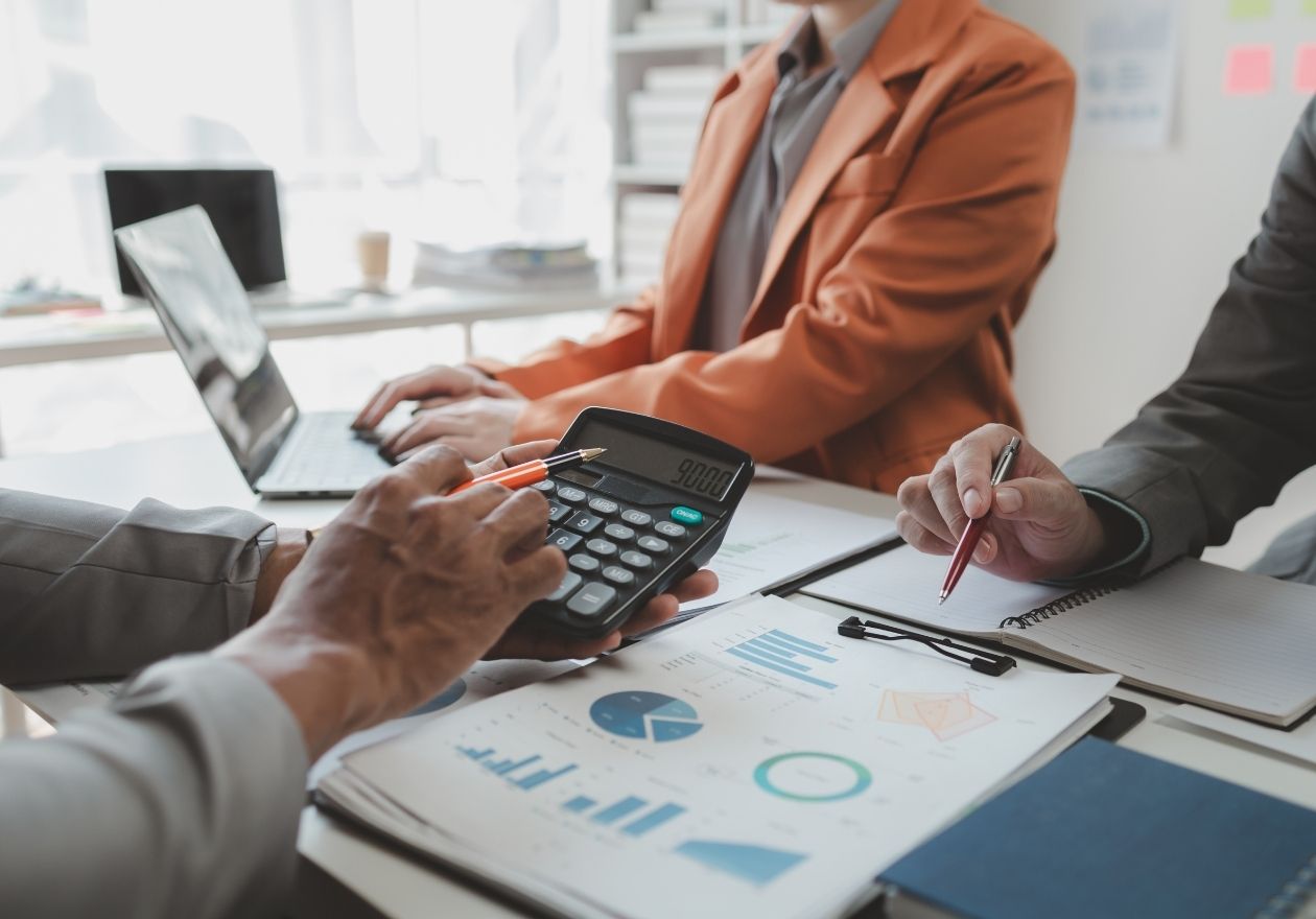 Pessoas em reunião analisam gráficos e usam calculadora sobre uma mesa com laptop. Curso: Aspectos Contábeis e Societários de uma Cooperativa.