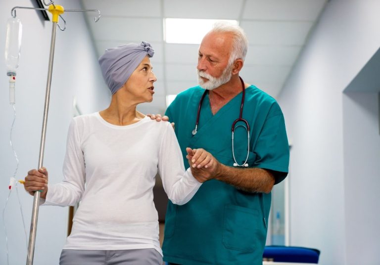 Médico apoia paciente com lenço na cabeça em um corredor hospitalar. Curso: Assistência ao Paciente Oncológico.
