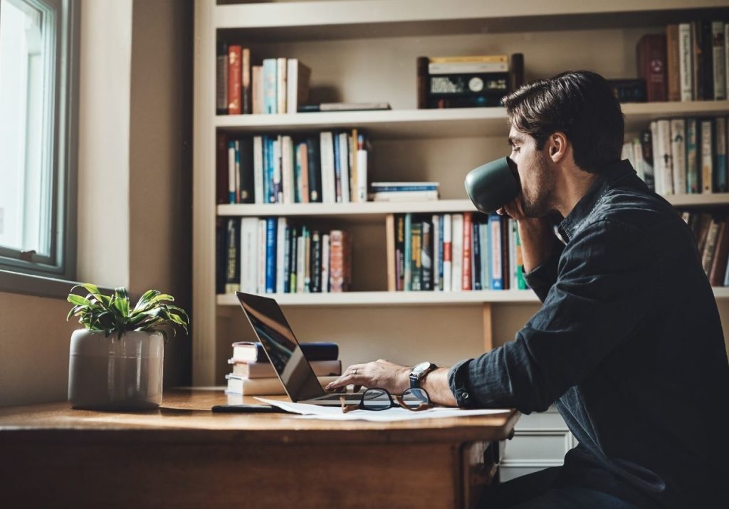 Homem trabalha em laptop e toma café. No fundo, uma estante com vários livros. Curso: Balancear a Vida.