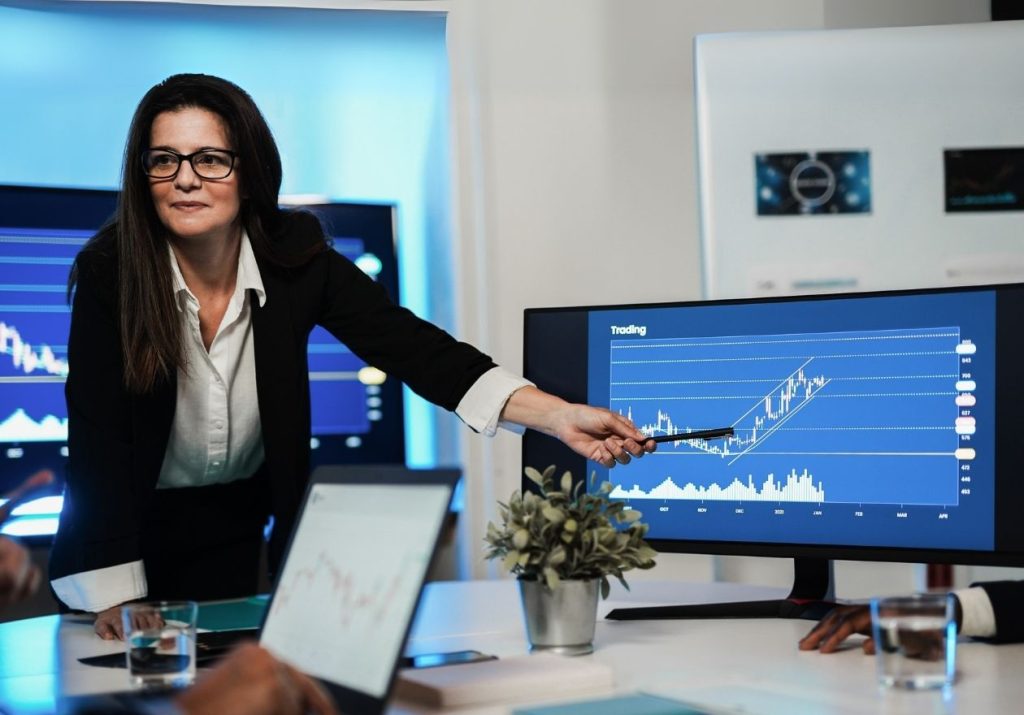 Mulher apresenta gráficos em tela, em sala com laptop e planta. Curso: Compreender a Economia.