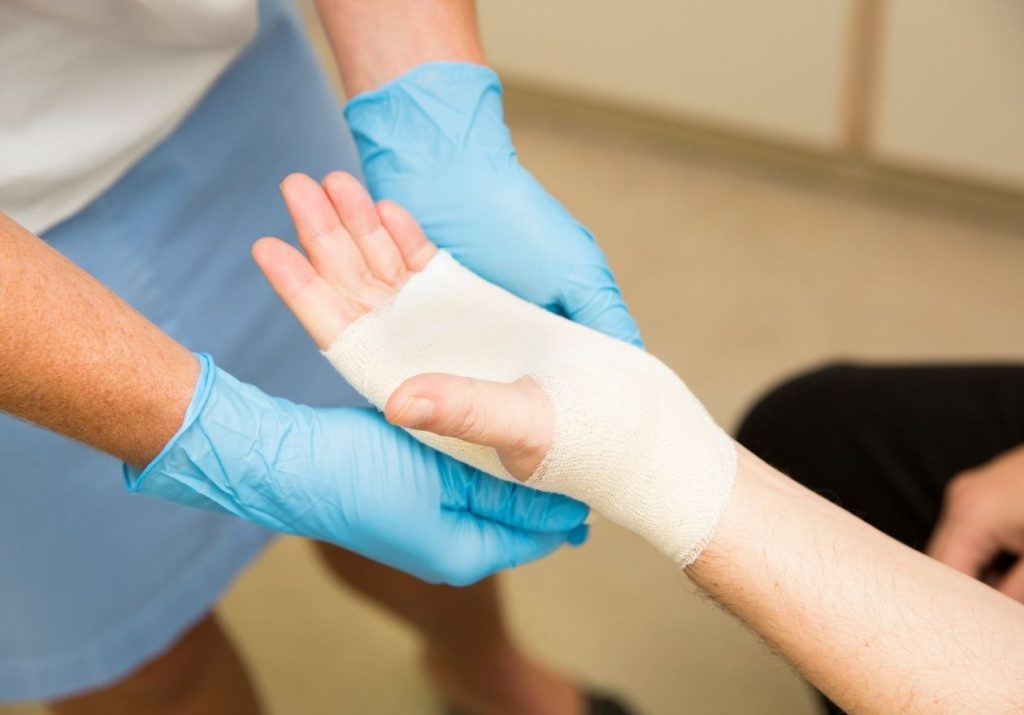 Um profissional de saúde com luvas azuis aplica uma bandagem na mão de um paciente. Curso: Condutas de Enfermagem no Tratamento de Feridas.
