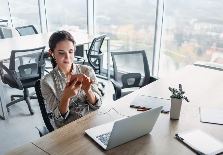 Uma mulher usa o celular em um escritório com laptop, planta e vista ampla pela janela. Curso: Criar uma Rotina Produtiva.
