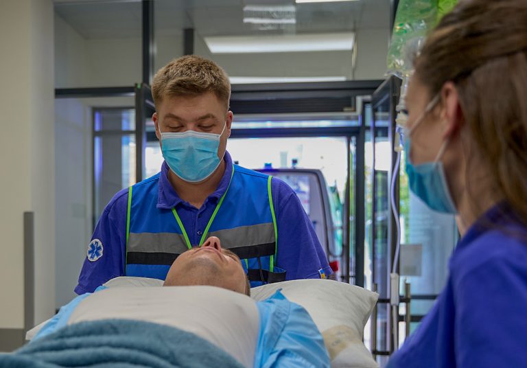 Dois profissionais de saúde atendem um paciente em uma maca. Eles estão na entrada de um hospital, com uma ambulância ao fundo. Curso: Enfermagem em Urgência, Emergência e Atendimento Pré-hospitalar.