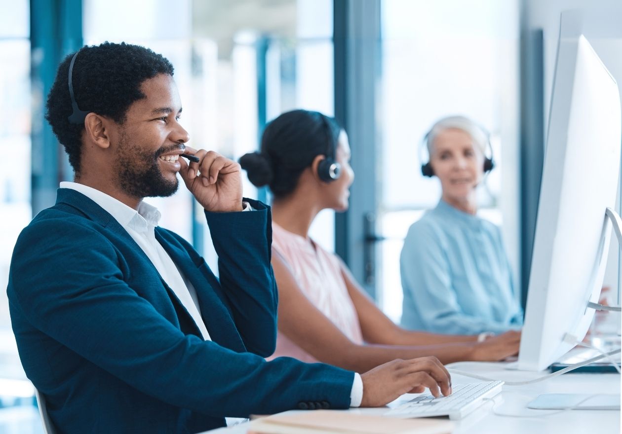 Três pessoas com headsets trabalham em computadores em um escritório, uma delas sorri ao telefone. Curso: Experiência do Cliente.