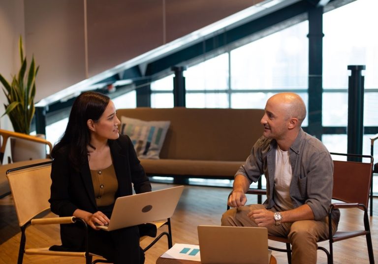 Duas pessoas conversam em um escritório com laptops, sentadas em cadeiras, com uma planta ao fundo. Curso: Feedforward.