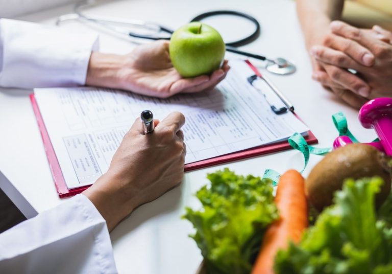 Profissional com estetoscópio segura uma maçã verde e analisa prancheta, com vegetais sobre a mesa. Curso: Gestão de Nutrição.