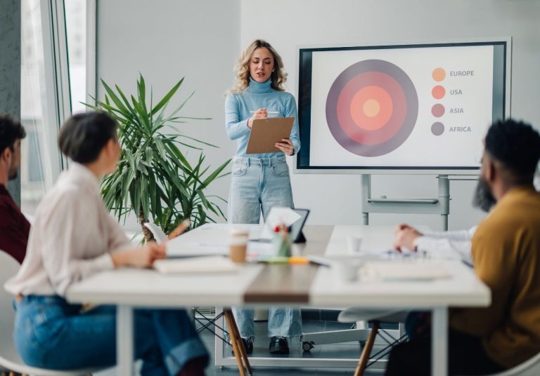 Mulher apresenta gráfico em tela para sua equipe. Curso: Gestão de Projetos.