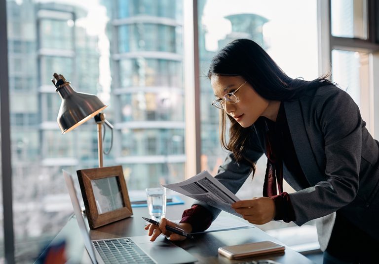 Mulher revisando documentos sobre gestão e planejamento financeiro para profissionais e clínicas de saúde em seu escritório, com notebook e luminária sobre a mesa em um ambiente profissional com vista para a cidade.