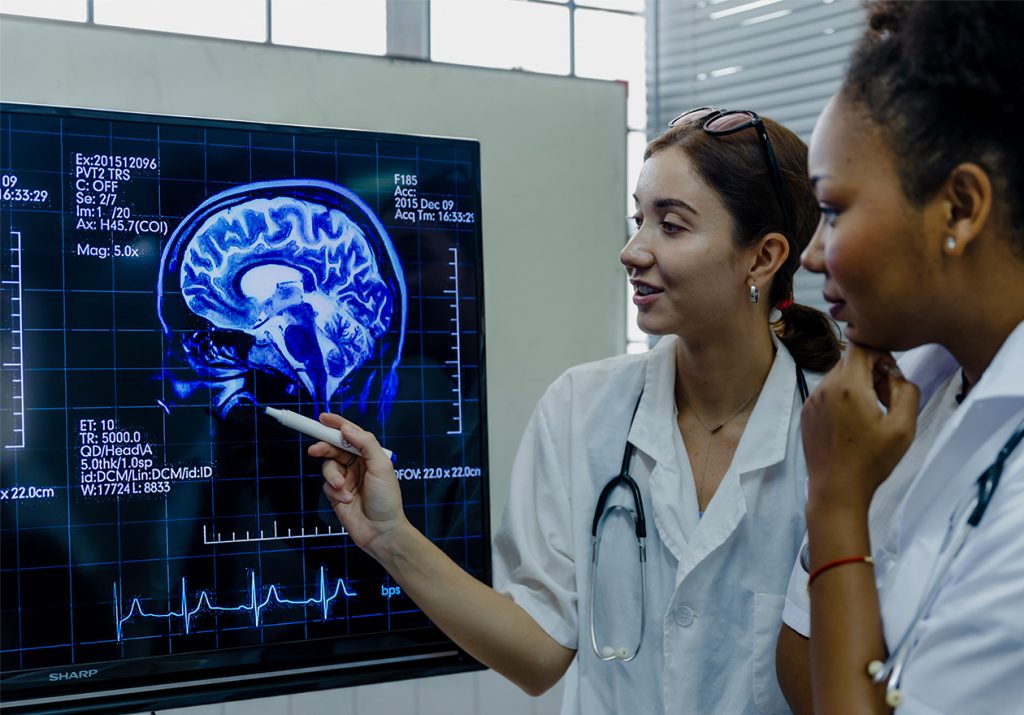 Duas médicas de jaleco analisam uma imagem de ressonância magnética do cérebro em uma tela. Curso: Neurociências e Comportamento.