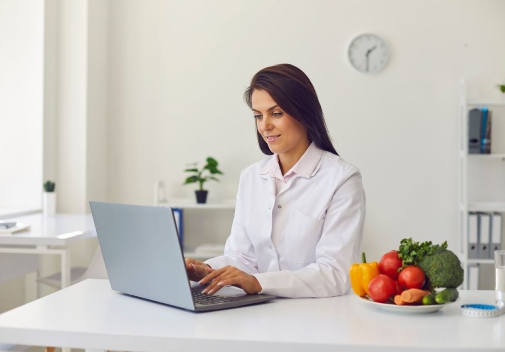 Uma profissional de jaleco trabalha em um laptop, com frutas e vegetais coloridos ao lado. Curso: Nutrição de Produção e Clínica.