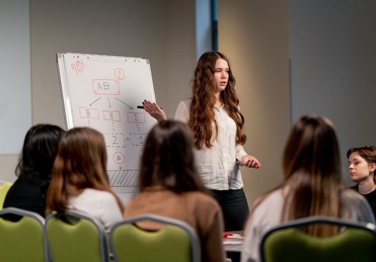 Mulher apresentando ideia em quadro para grupo sentado em sala, com cadeiras verdes ao redor. Curso: Vender uma Ideia em 60 Segundos.