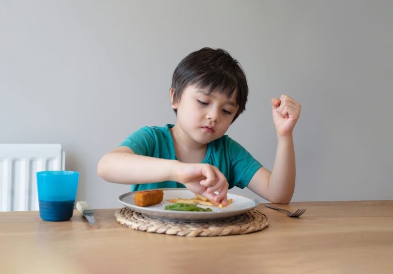Criança sentada à mesa mexe em comida no prato, com copo e talheres ao lado. Curso: Capacitação em Seletividade Alimentar.