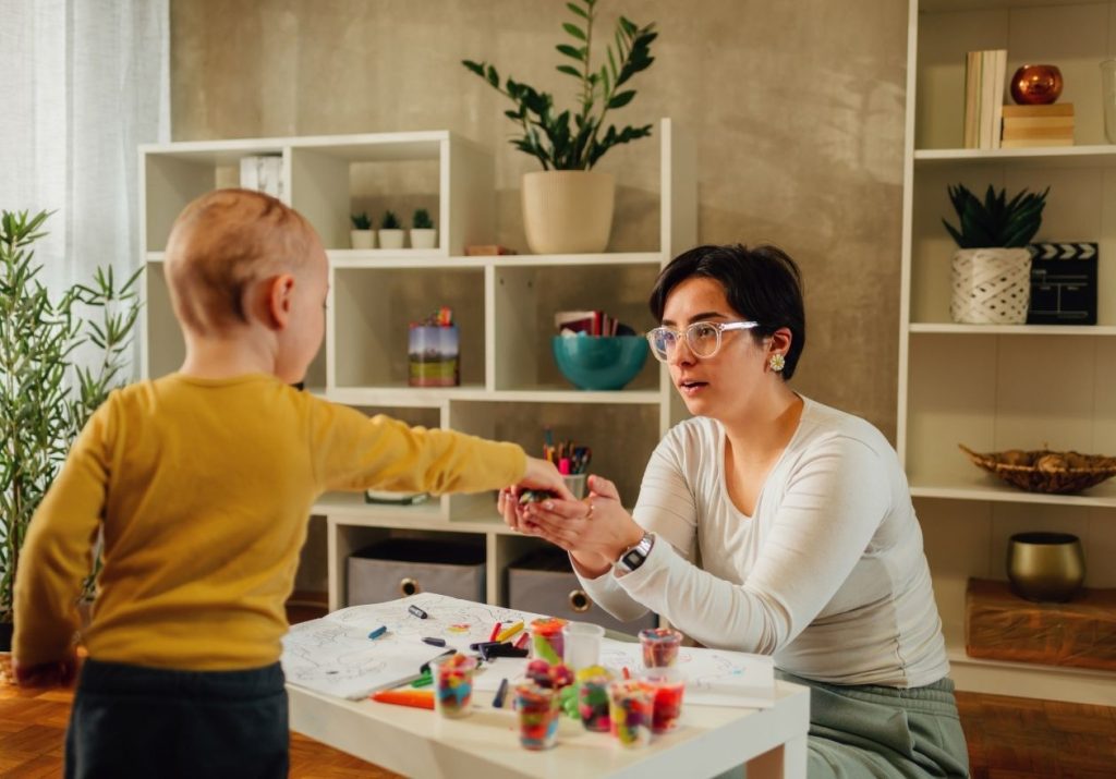 Mulher e criança interagem com objetos coloridos em mesa, em sala com plantas e estantes. Curso: Capacitação em VBMAPP e Programação Terapêutica.