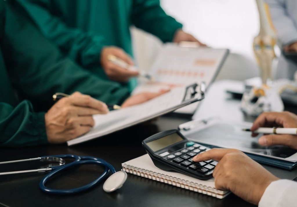 Profissionais de saúde analisam papéis e usam calculadora, com estetoscópio na mesa. Curso: Finanças Pessoais para Médicos.