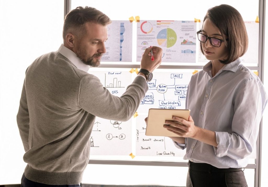 Homem e mulher conversam em frente a um quadro com gráficos e anotações. A mulher está segurando um tablet. Curso: Gestão Administrativa e Financeira.