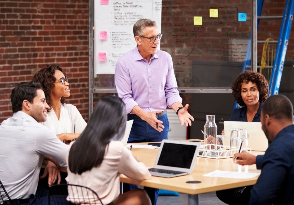 Homem conversa com equipe em reunião, quadro de anotações ao fundo. Curso: Gestão da Mudança.