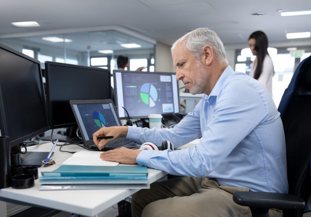 Homem trabalha em estação com múltiplos monitores exibindo gráficos. Curso: Gestão de Tecnologia e Segurança da Informação.