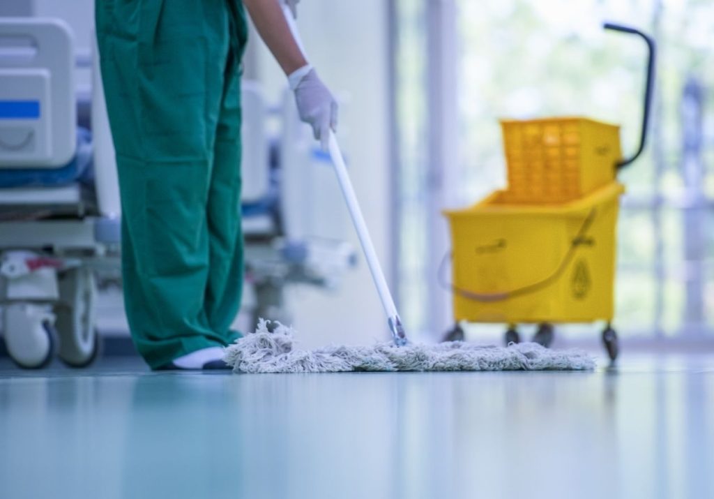 Profissional limpa chão de hospital com esfregão, balde e equipamento médico ao fundo. Curso: Higienização.