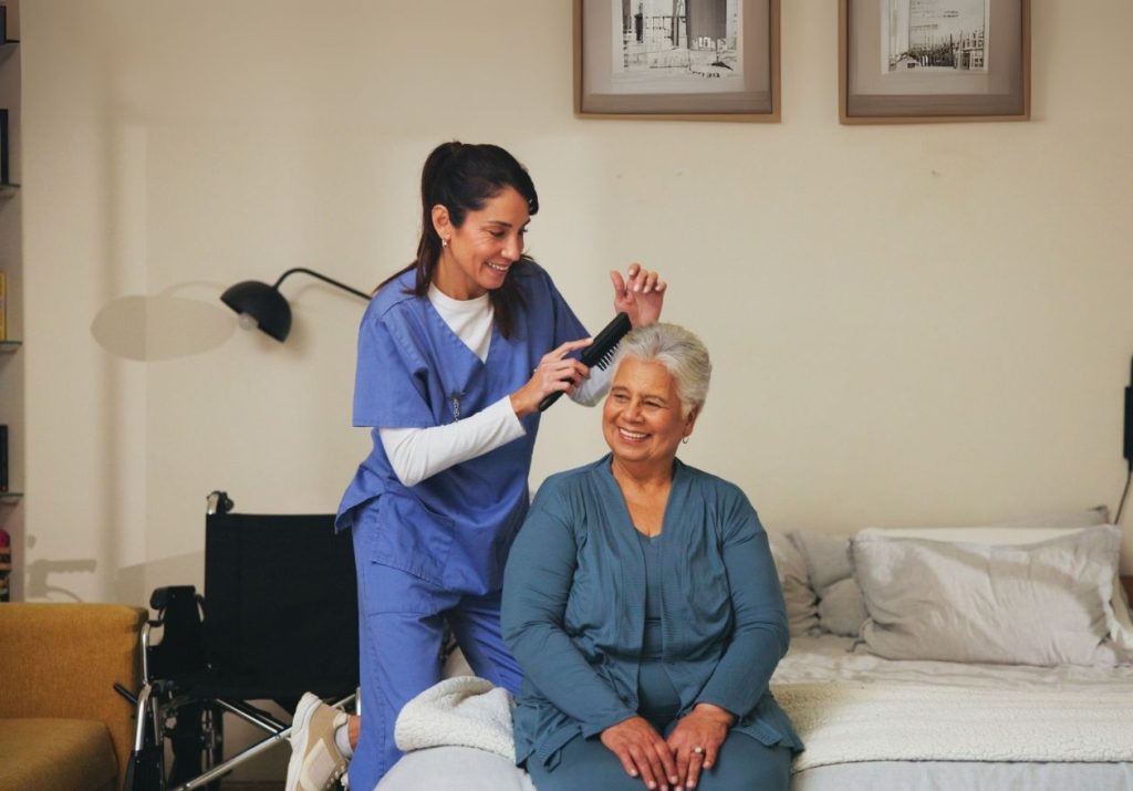 Cuidadora penteia cabelo de idosa sentada em cama. Elas estão em um quarto com quadros e cadeira de rodas. Curso: Incapacidade Cognitiva e Alzheimer.