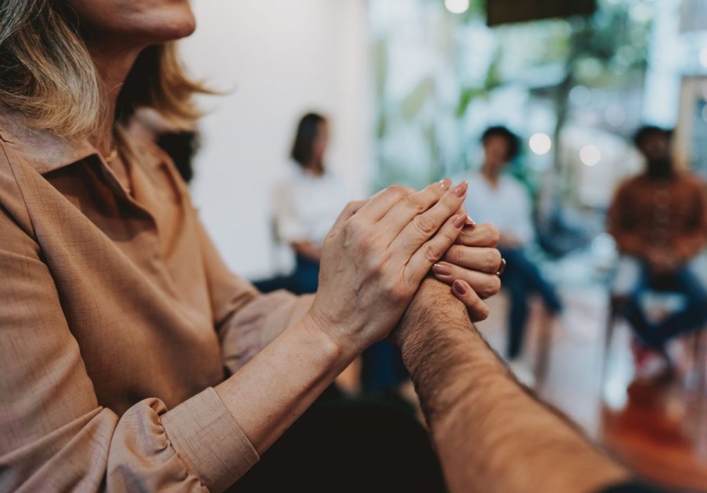 Pessoas seguram as mãos em gesto de apoio. Outras pessoas estão sentadas ao fundo. Curso: Inteligência Emocional.