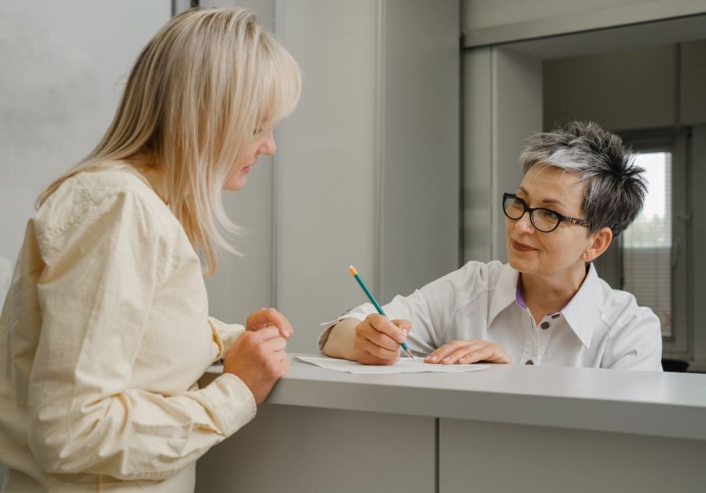 Mulher conversa com atendente em balcão, que anota com lápis em papel. Curso: Intercâmbio.