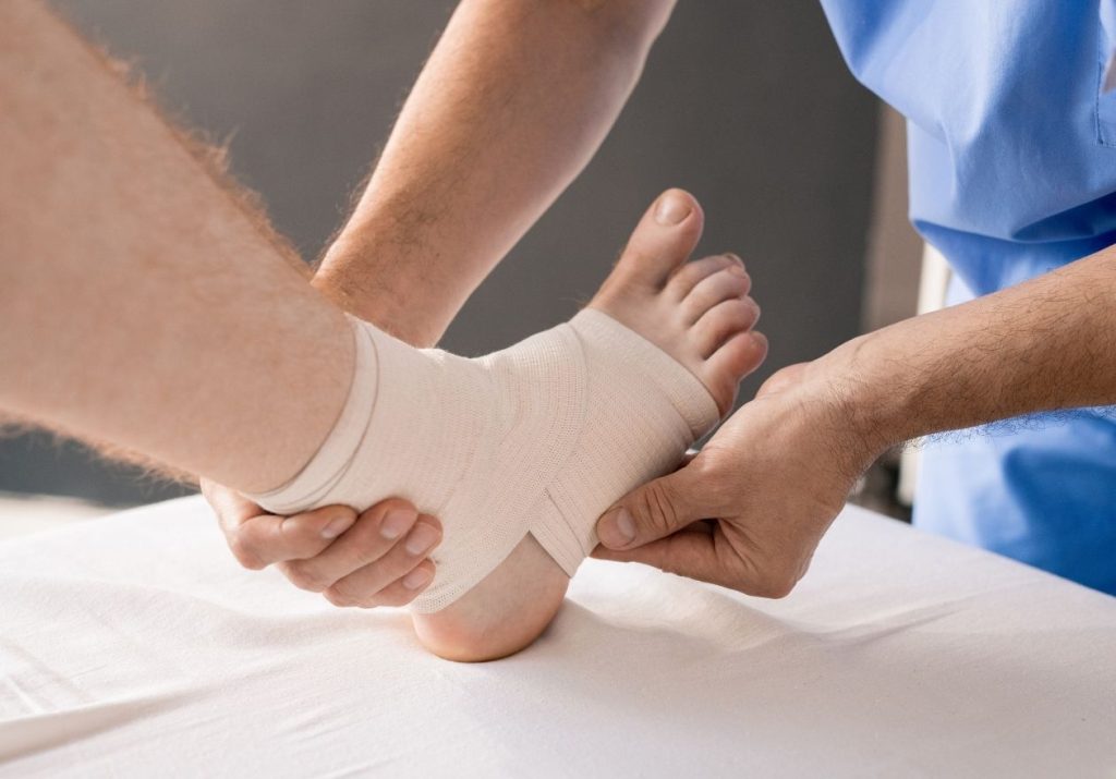 Profissional da saúde aplica uma bandagem, tratando uma lesão em ambiente hospitalar. Curso: Lesões Ligamentares.
