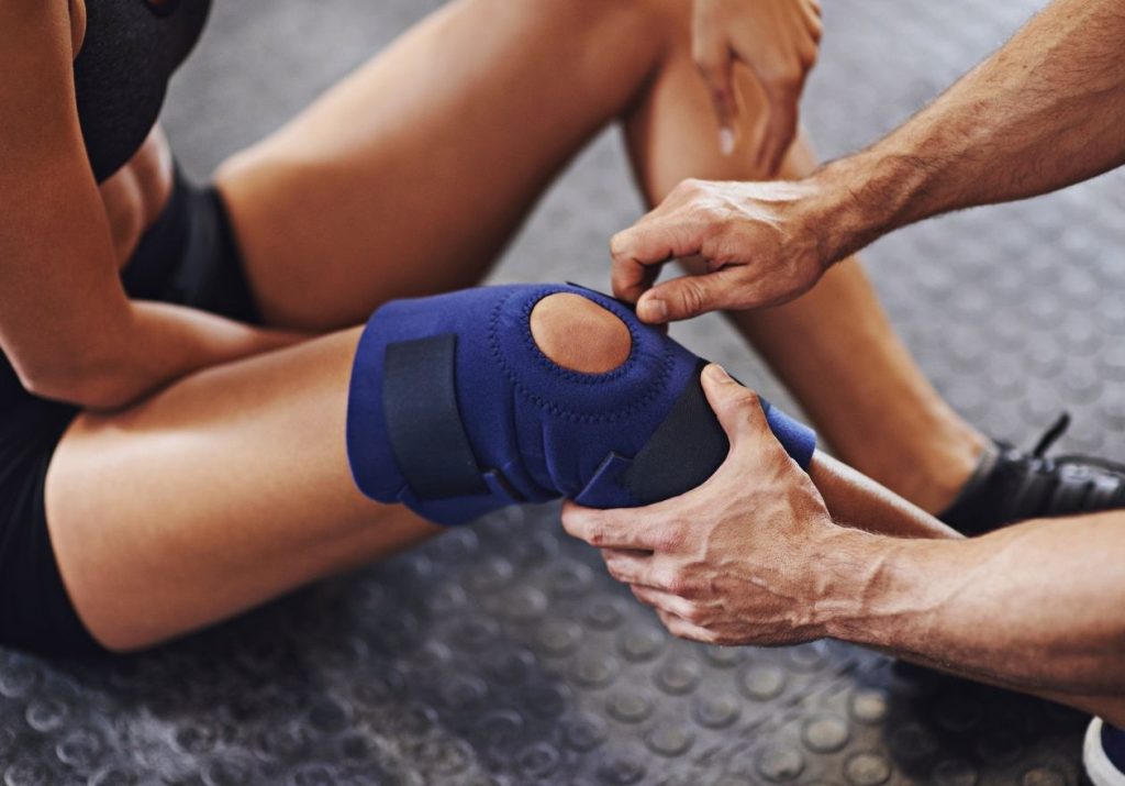 Profissional ajusta uma joelheira azul no joelho de uma mulher, em um ambiente de reabilitação ou treino. Curso: Lesões Meniscais.