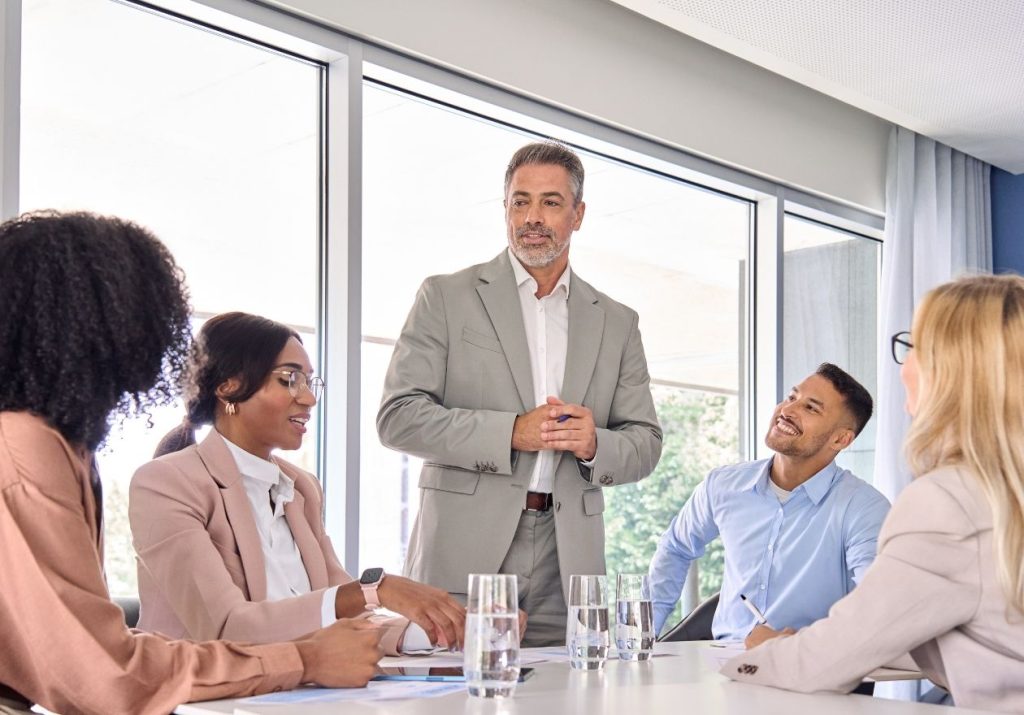 Homem de terno fala para grupo sentado ao redor de uma mesa com copos d'água, em sala iluminada. Curso: Liderança Adaptativa.