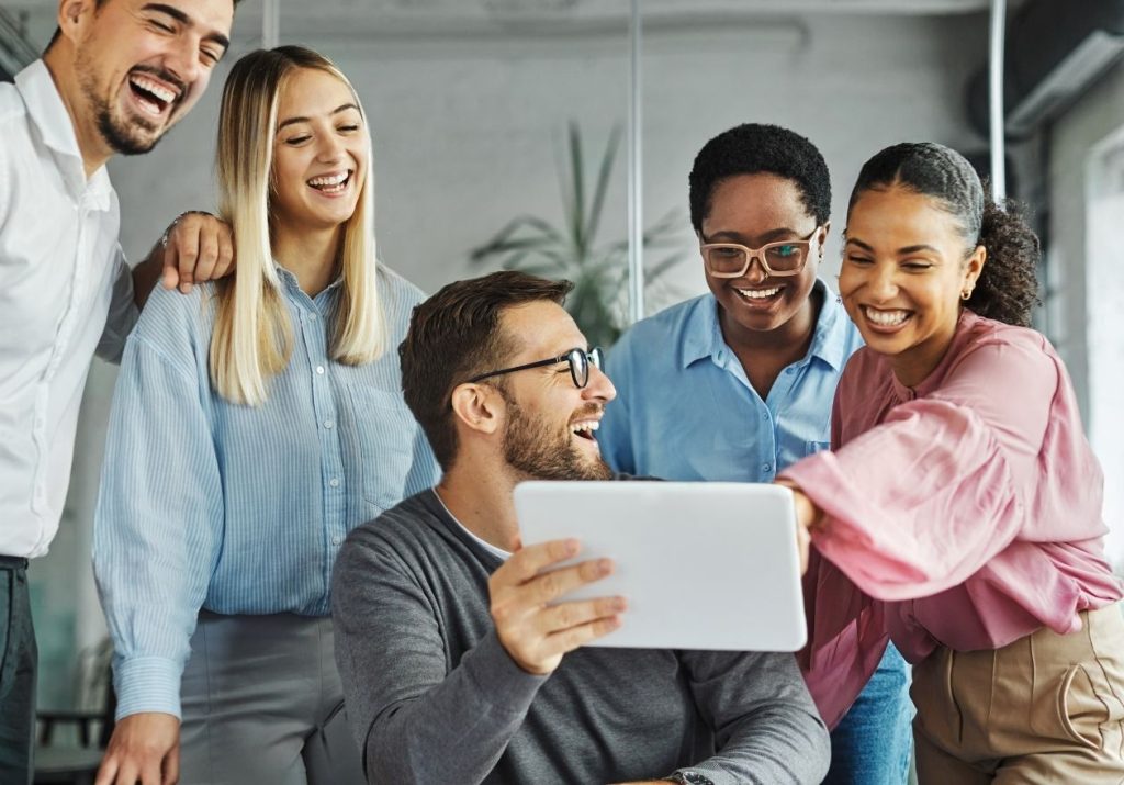 Grupo de cinco pessoas sorrindo e interagindo com um tablet em ambiente descontraído com planta ao fundo. Curso: Liderança Carismática.