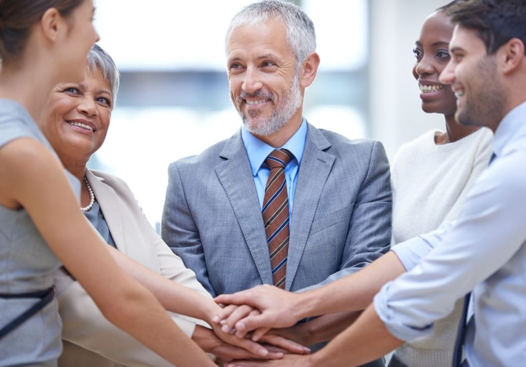 Cinco pessoas unem as mãos em círculo, sorrindo, em um ambiente profissional. Curso: Liderança e Desenvolvimento de Equipes em Cooperativas.