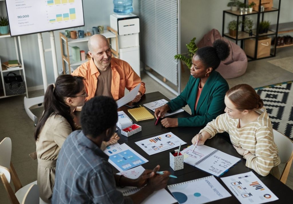 Cinco pessoas discutem ao redor de uma mesa com gráficos e documentos, em sala com quadro e plantas. Curso: Liderança e Gestão.