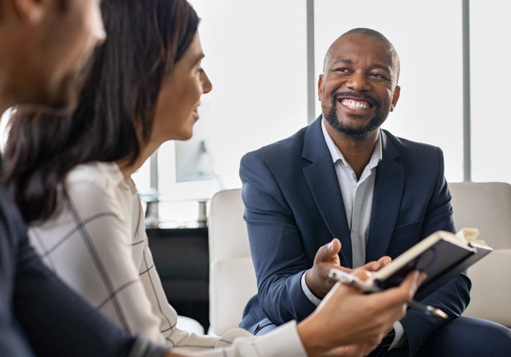 Três pessoas conversam em um ambiente profissional, uma delas segurando um caderno, discutindo estratégias. Curso: Melhorar a Prospecção em Vendas.
