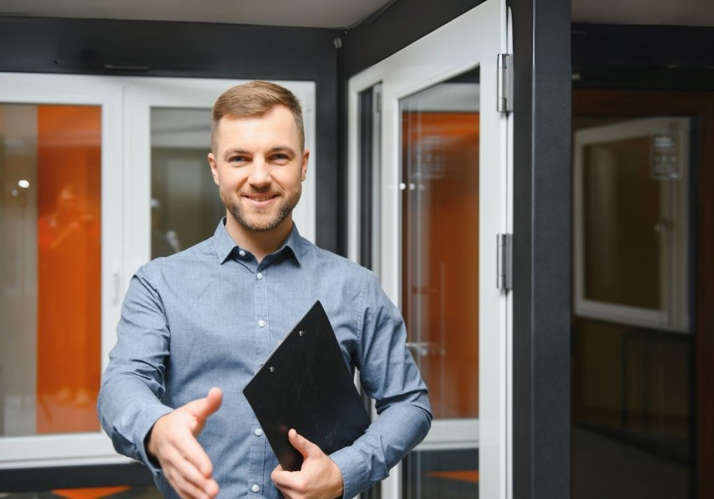 Homem sorridente em um escritório segura uma prancheta e oferece um aperto de mão. Curso: Melhorar as Técnicas de Fechamento de Vendas.