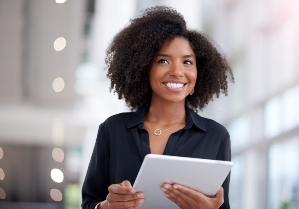 Mulher sorridente segura um tablet em um ambiente de escritório moderno com grandes janelas e luz natural. Curso: Mentes Poderosas.