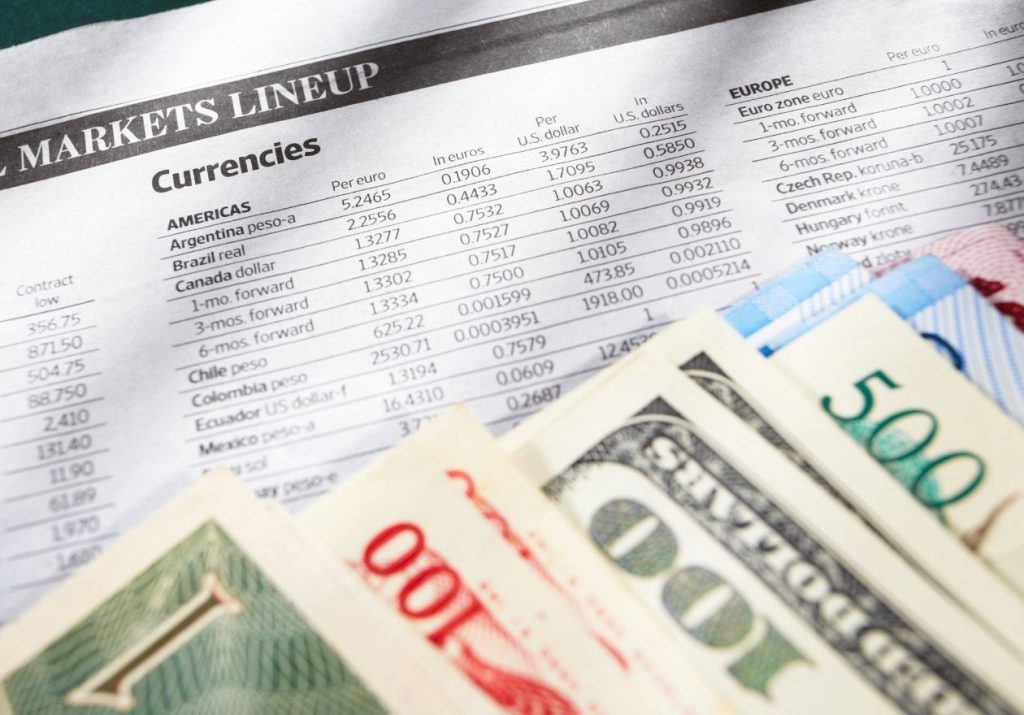 Notas de diferentes moedas e um jornal financeiro sobre uma mesa. Curso: Mercado de Câmbio - Abordagem Estratégica.