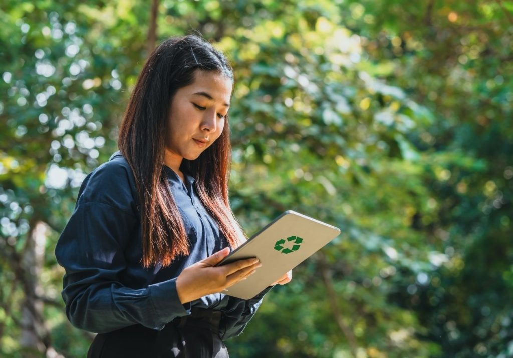 Uma mulher segura um tablet com símbolo de reciclagem em um ambiente natural com árvores ao fundo. Curso: Mercado de Crédito de Carbono.