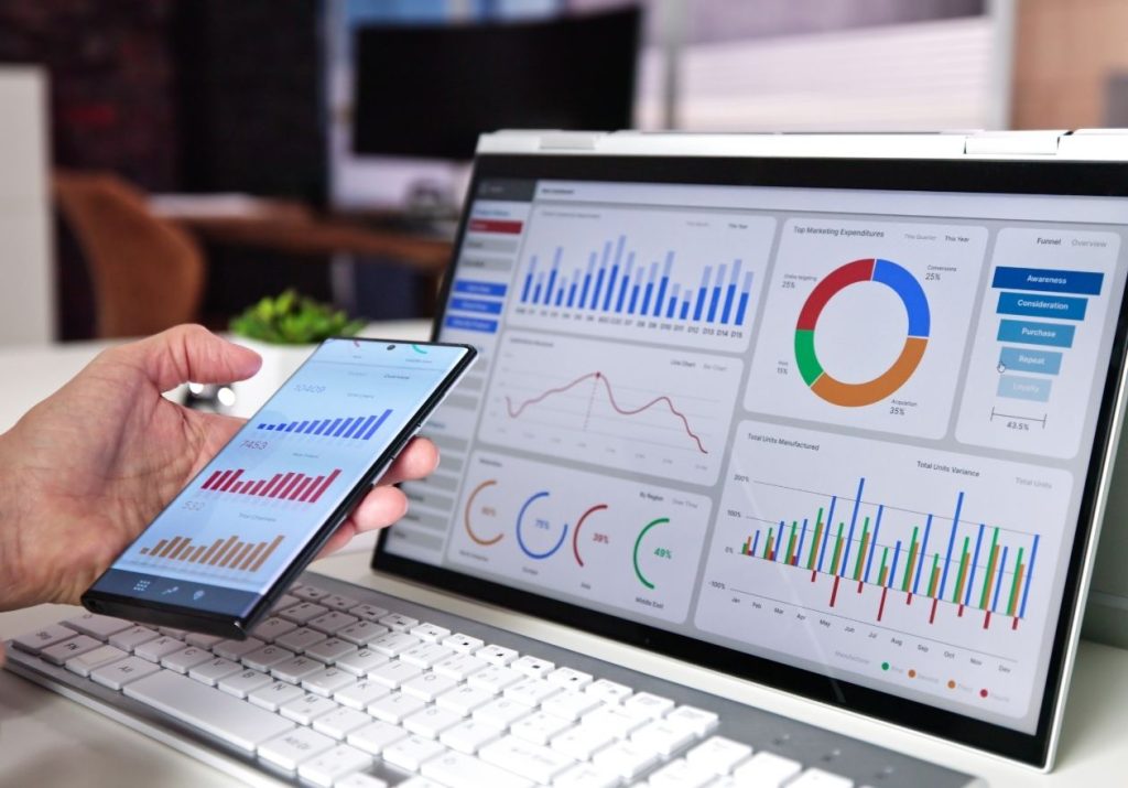 Uma mão segura um celular ao lado de um monitor exibindo gráficos e métricas coloridas. Curso: Métricas Ágeis.